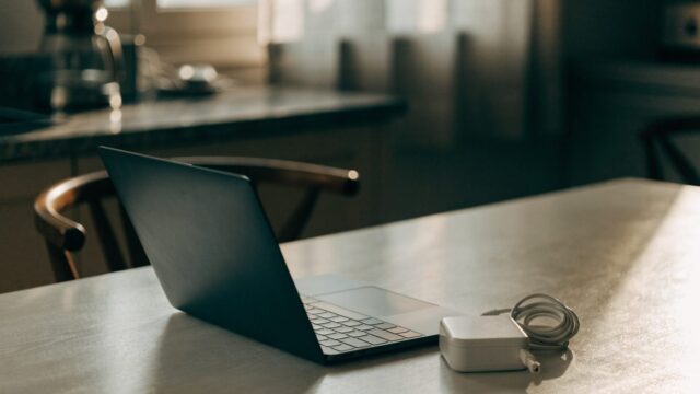 A graphite black laptop on a kitchen table next to a compact white power adapter with its cable neatly coiled