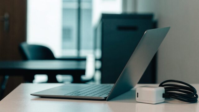 A matte grey laptop on an office desk next to a compact white power adapter with its cable neatly coiled