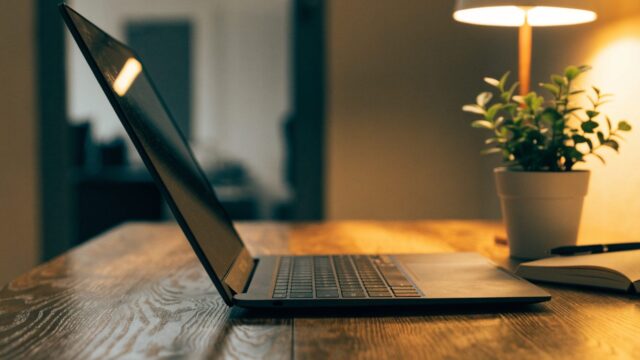A graphite black laptop open on a wooden desk with a small plant and notebook beside it