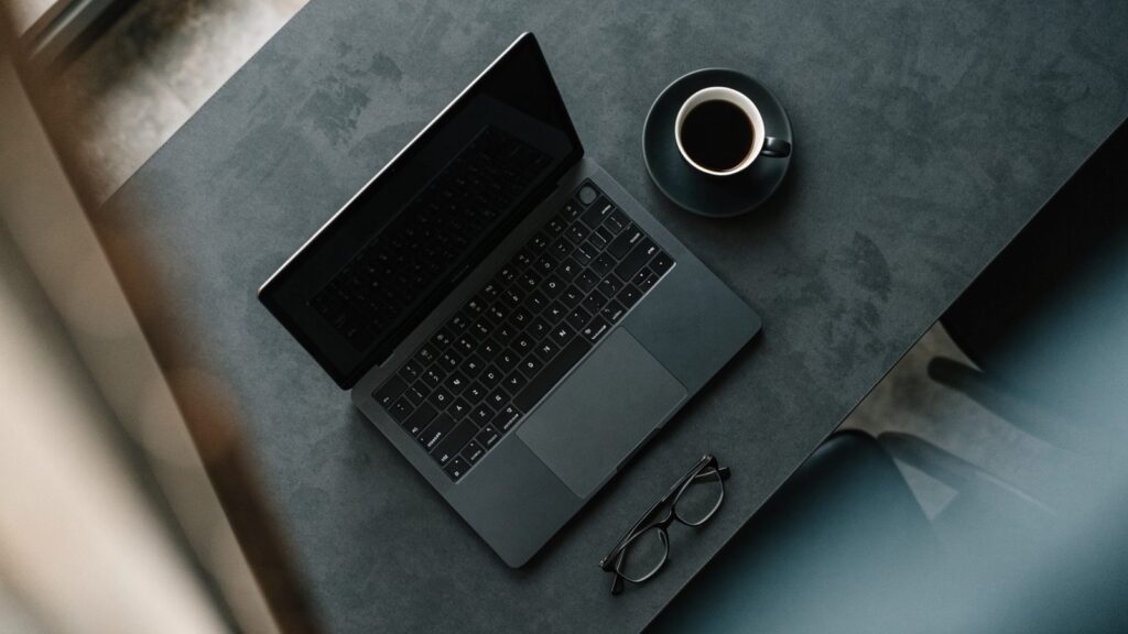Top-down view of a graphite black laptop on a dark desk mat with a coffee cup and glasses