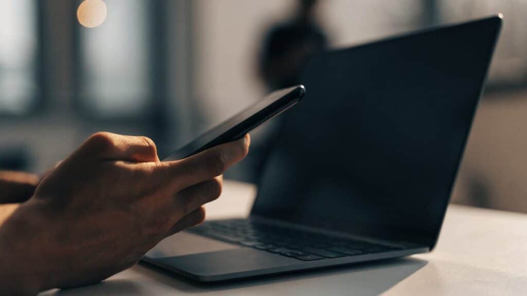 A hand holding a smartphone in front of a matte grey laptop on a white desk