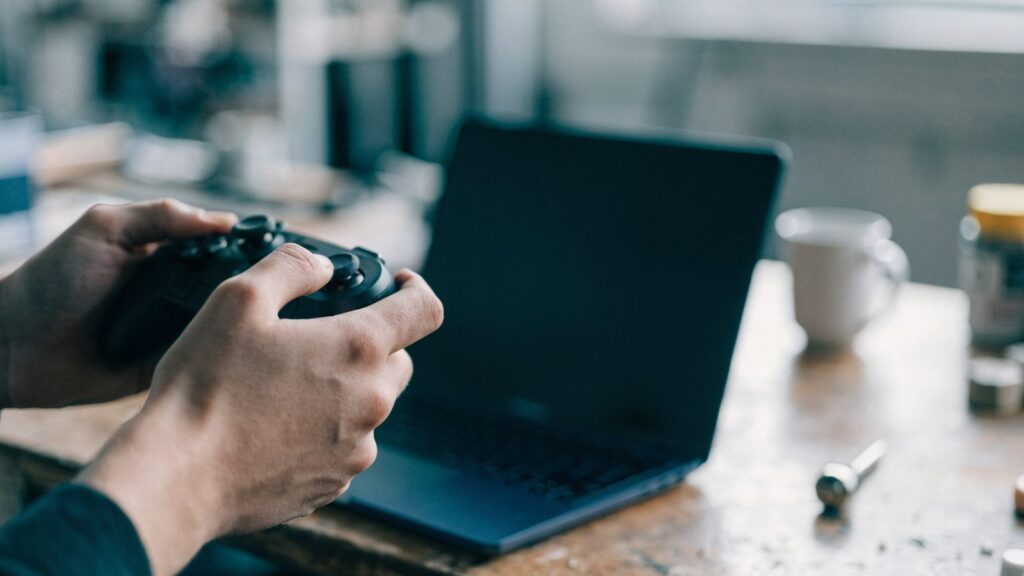 Hands holding a game controller in front of a dark blue laptop on a workbench