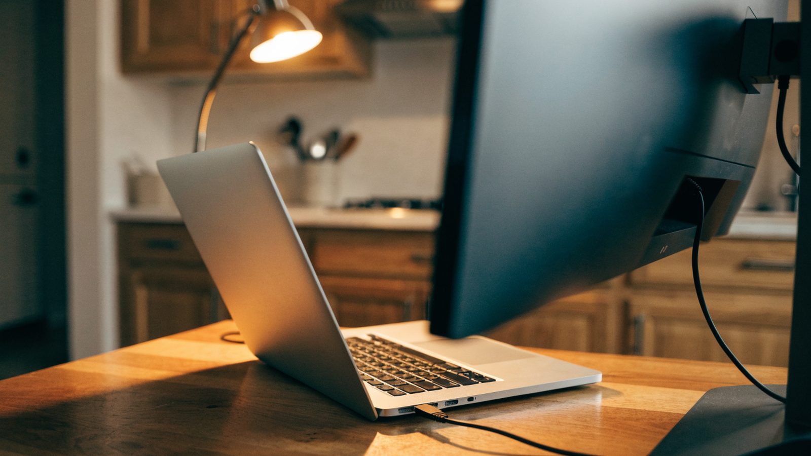 A silver laptop connected to a large external monitor with a blank dark screen via a cable on a kitchen table