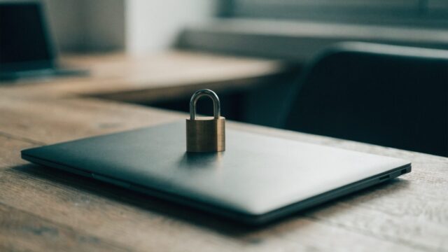 A small brass padlock resting on the closed lid of a graphite black laptop on a wooden desk