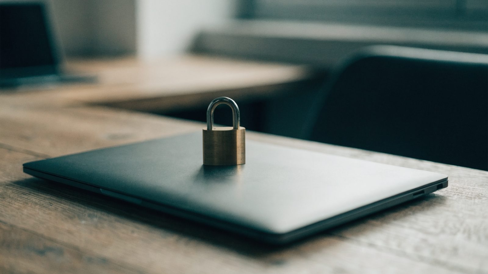A small brass padlock resting on the closed lid of a graphite black laptop on a wooden desk