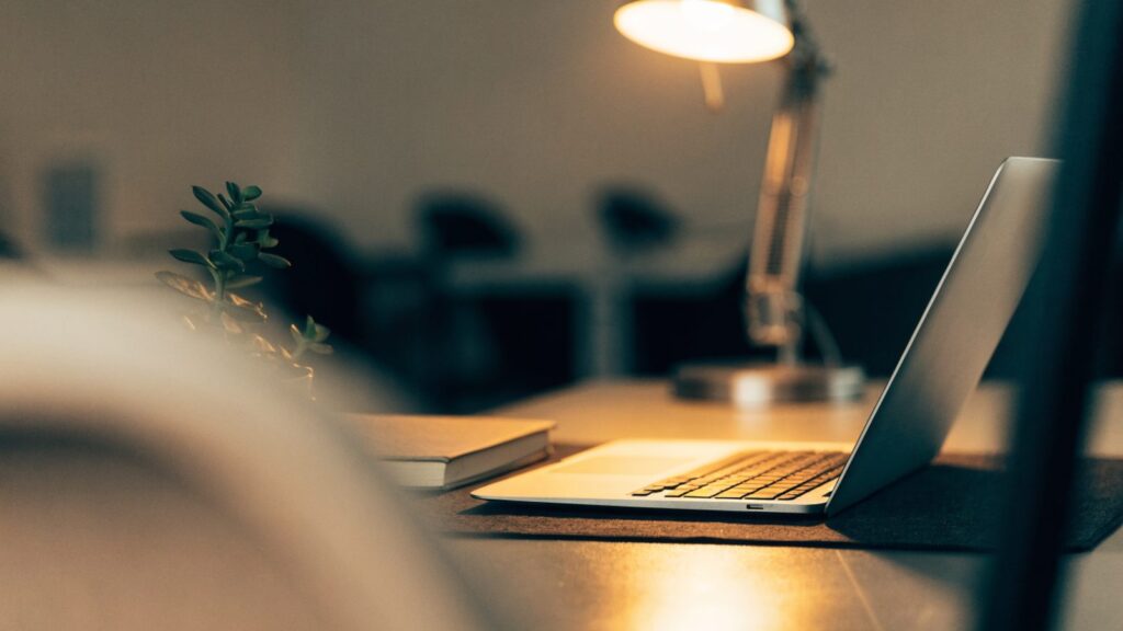 A silver laptop open on a dark desk mat with a small plant and notebook beside it