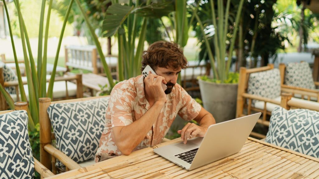 Handsome man with beard using lap top and mobyle phone sitting in outdoor cafe with tropical interior