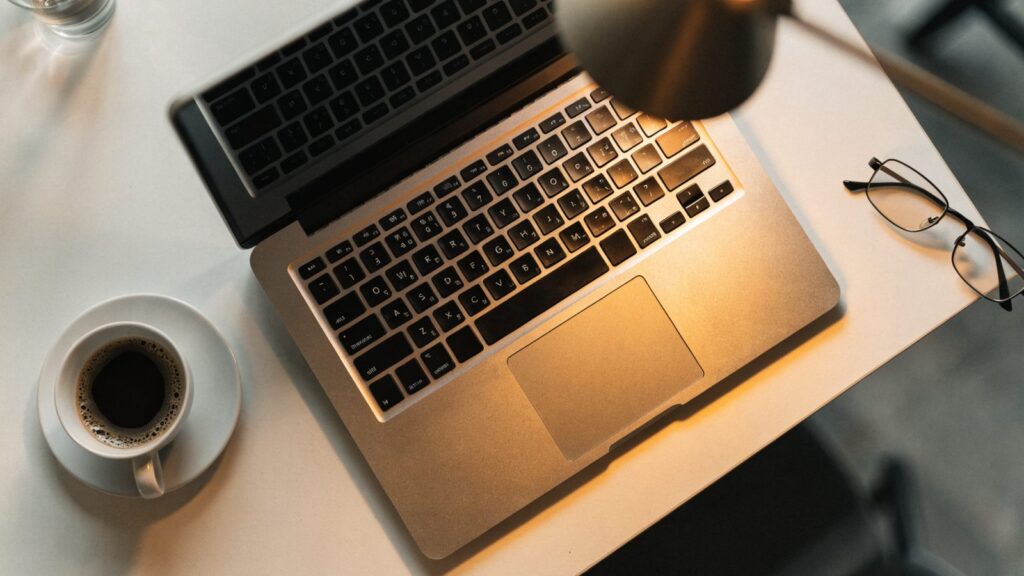Top-down view of a silver laptop on a white desk with a coffee cup and glasses