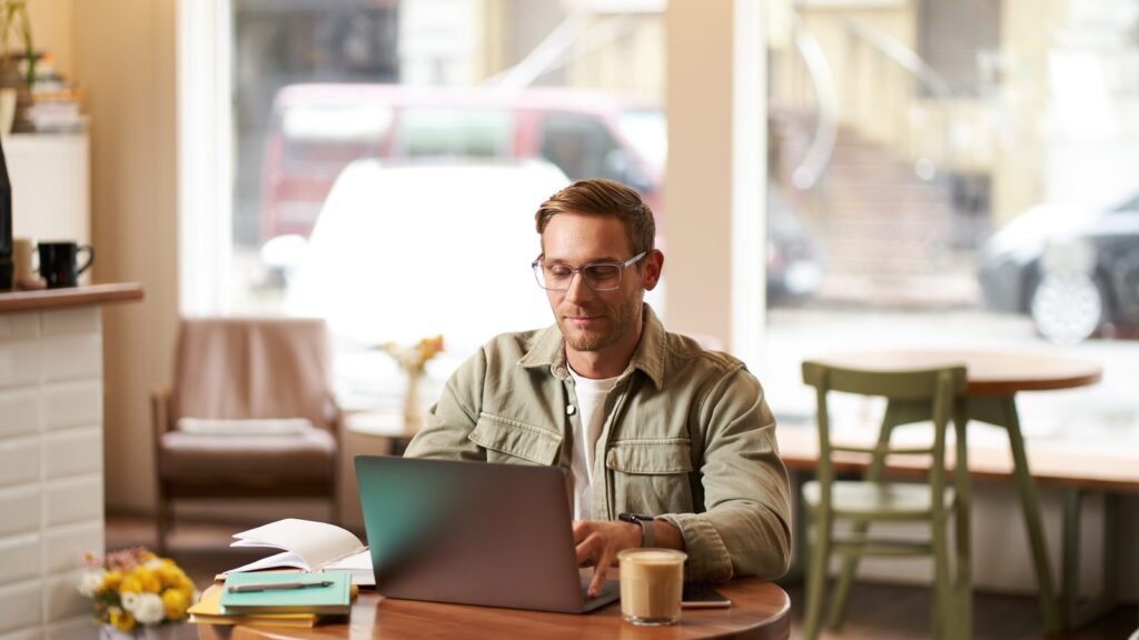 Image of young digital nomad man in glasses sits in cafe works from coffee shop uses laptop in