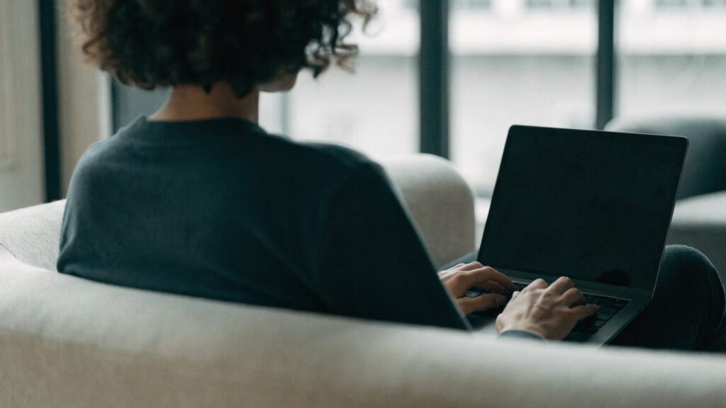 A person seen from behind using a graphite black laptop on a sofa