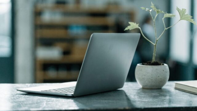 A matte grey laptop open on a workbench with a small plant and notebook beside it