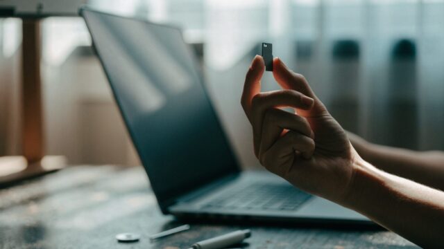 An SD card held between two fingers in front of a matte grey laptop on a workbench