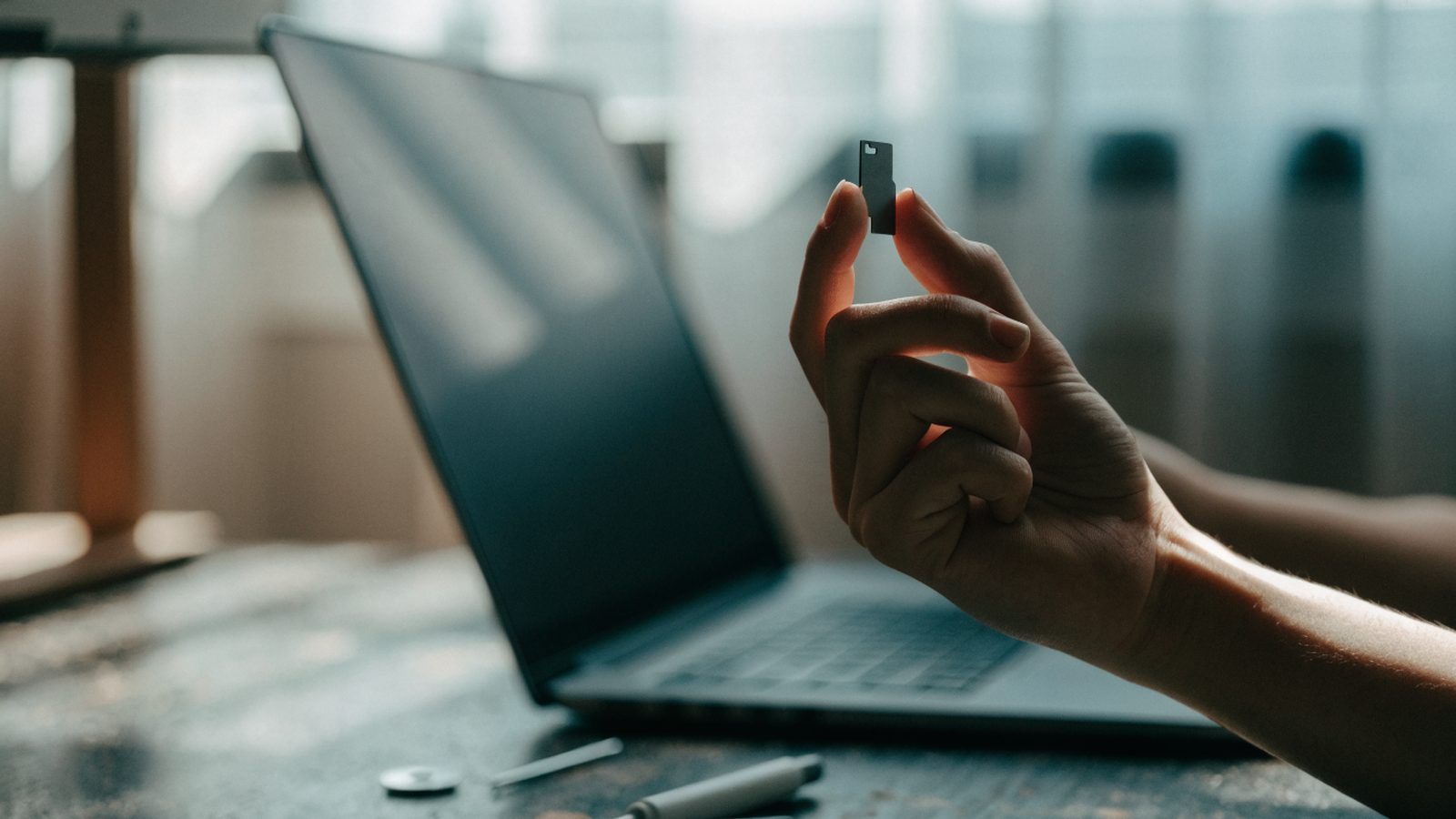 An SD card held between two fingers in front of a matte grey laptop on a workbench