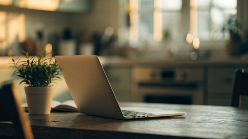 A silver laptop open on a kitchen table with a small plant and notebook beside it