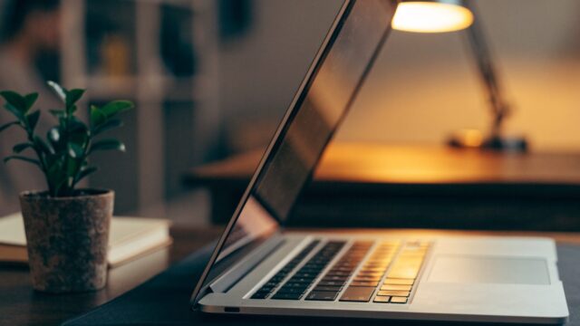 A silver laptop open on a dark desk mat with a small plant and notebook beside it