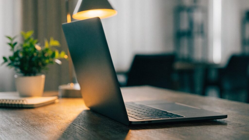 A graphite black laptop open on an office desk with a small plant and notebook beside it
