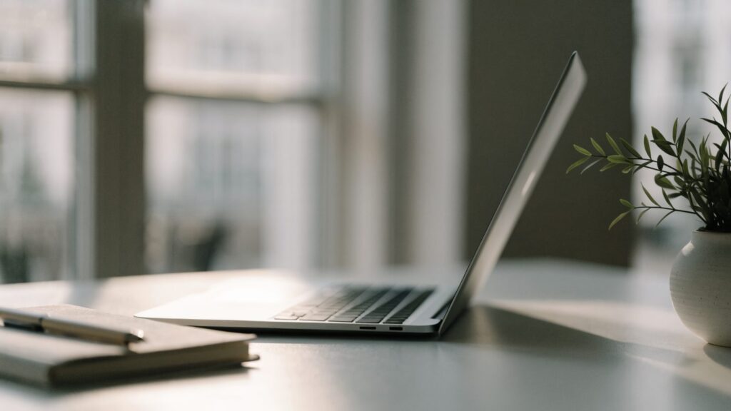 A silver laptop open on a white desk with a small plant and notebook beside it