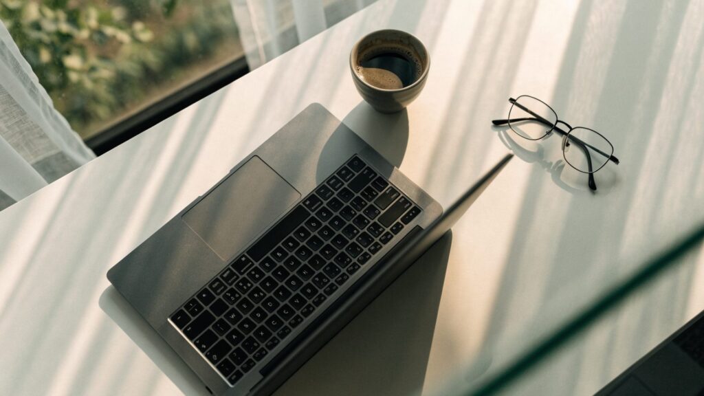 Top-down view of a matte grey laptop on a white desk with a coffee cup and glasses