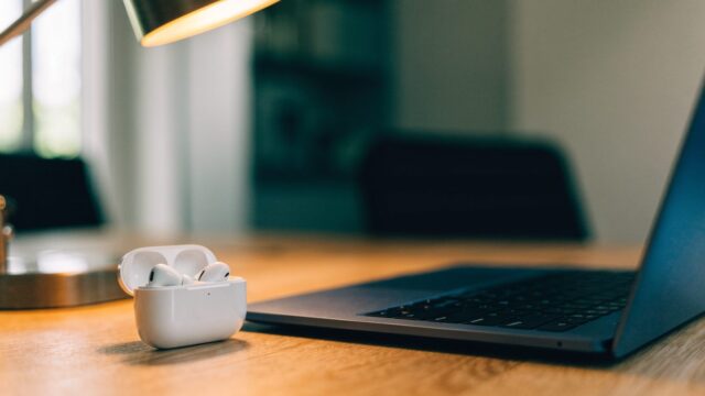 White wireless earbuds in their open case beside a dark blue laptop on an office desk