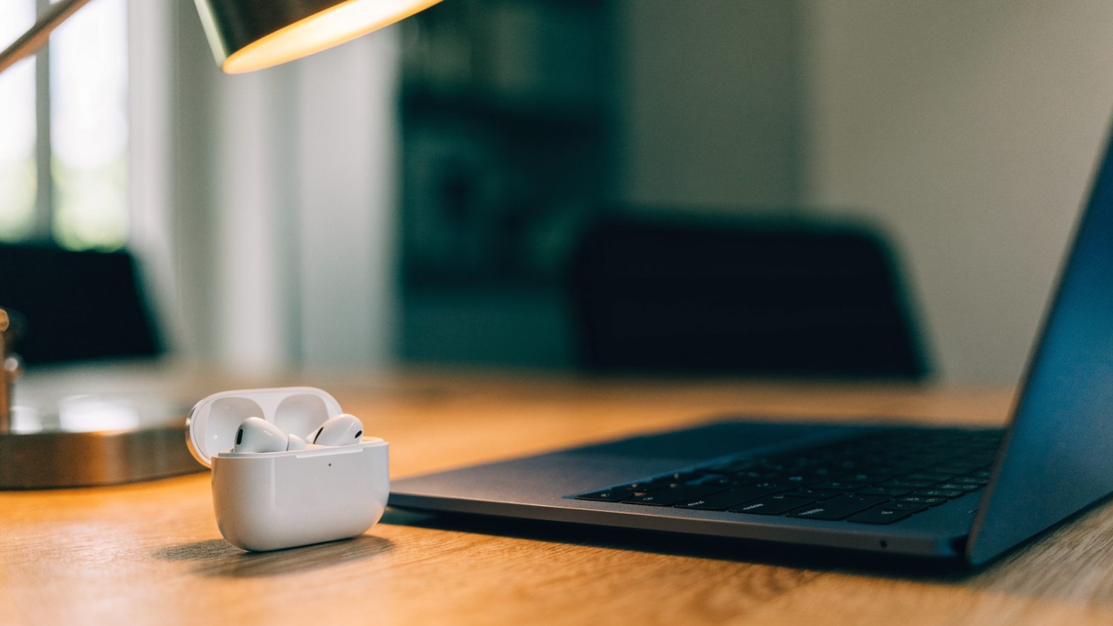 White wireless earbuds in their open case beside a dark blue laptop on an office desk
