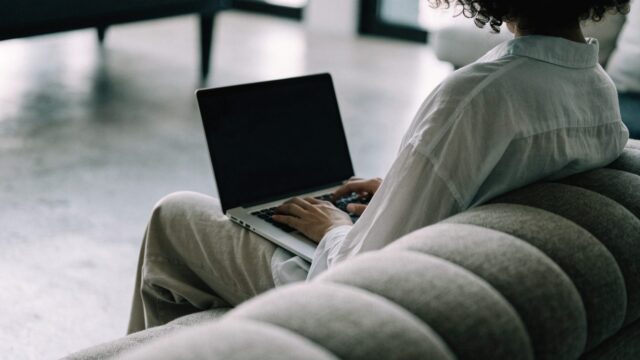 A person seen from behind using a silver laptop on a sofa