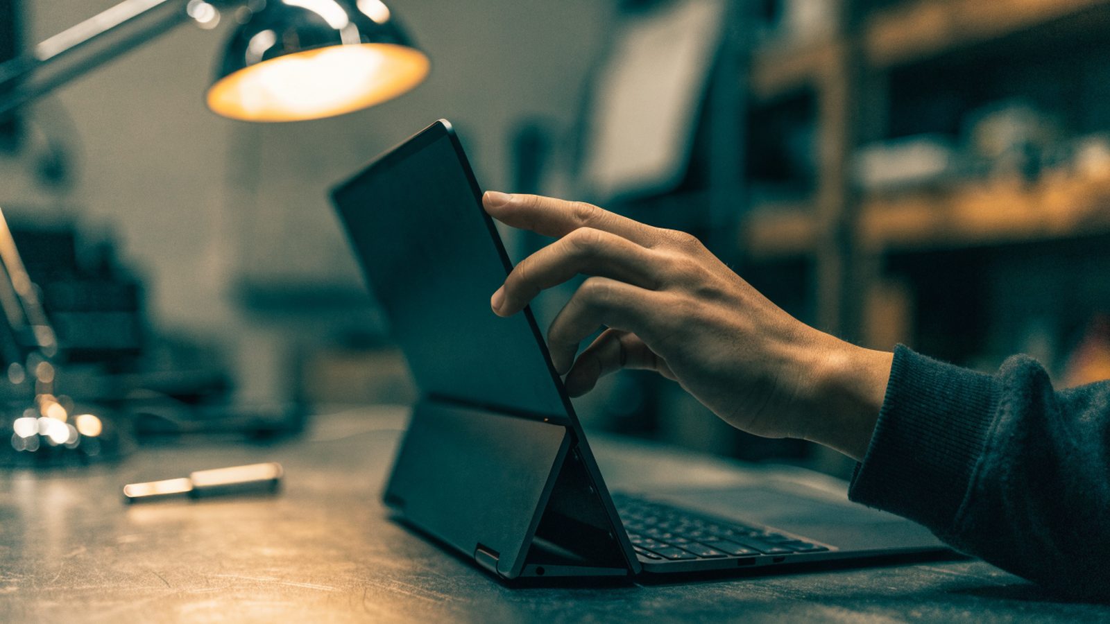 A hand touching the screen of a convertible graphite black laptop folded in tent mode on a workbench