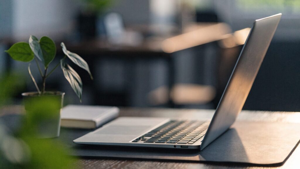 A silver laptop open on a dark desk mat with a small plant and notebook beside it