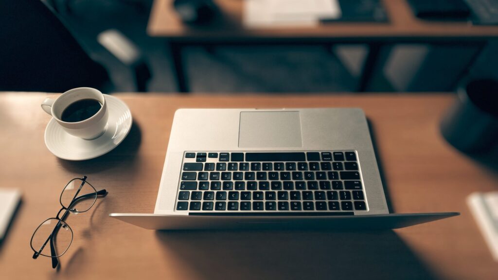 Top-down view of a silver laptop on an office desk with a coffee cup and glasses