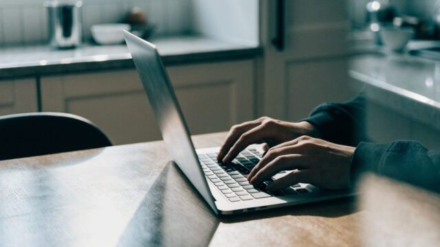 Hands typing on the keyboard of a silver laptop on a kitchen table