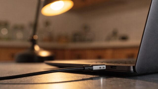 Close-up of a USB-C charger cable plugged into the side port of a graphite black laptop on a kitchen table