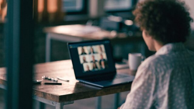 A person seen from behind on a video call on a dark blue laptop on a workbench