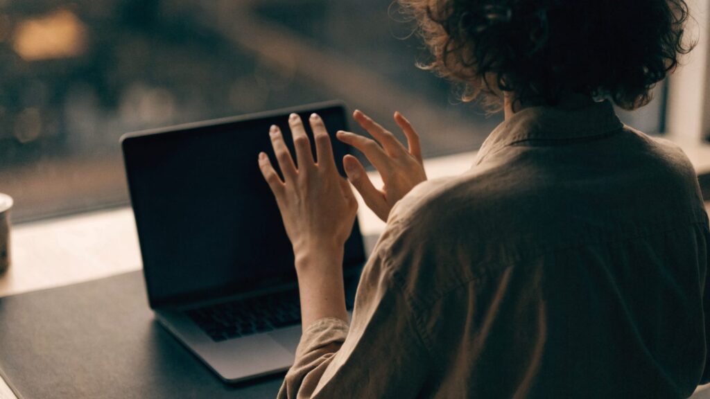A person seen from behind with hands raised at a matte grey laptop with a dark blank screen on a dark desk mat