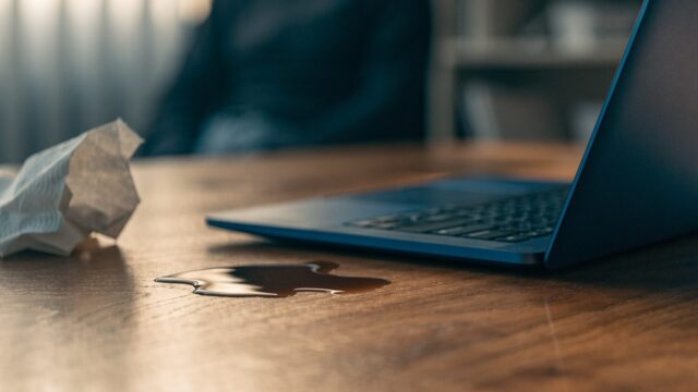 A small puddle of spilled coffee next to a dark blue laptop on a wooden desk