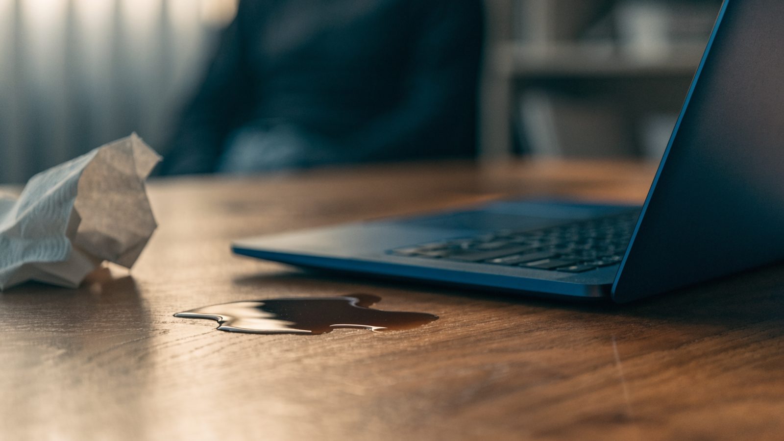 A small puddle of spilled coffee next to a dark blue laptop on a wooden desk