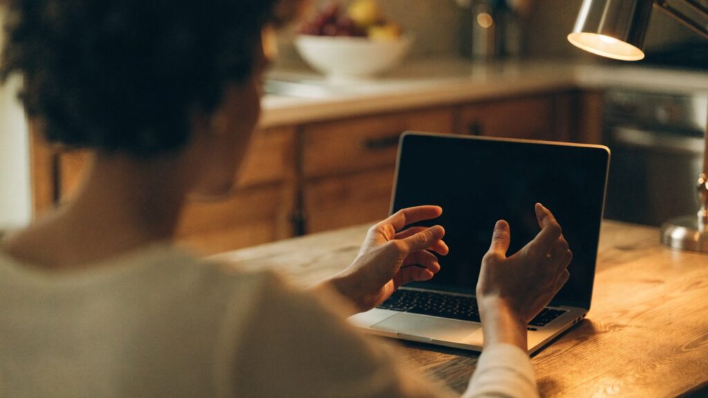 A person seen from behind with hands raised at a silver laptop with a dark blank screen on a kitchen table