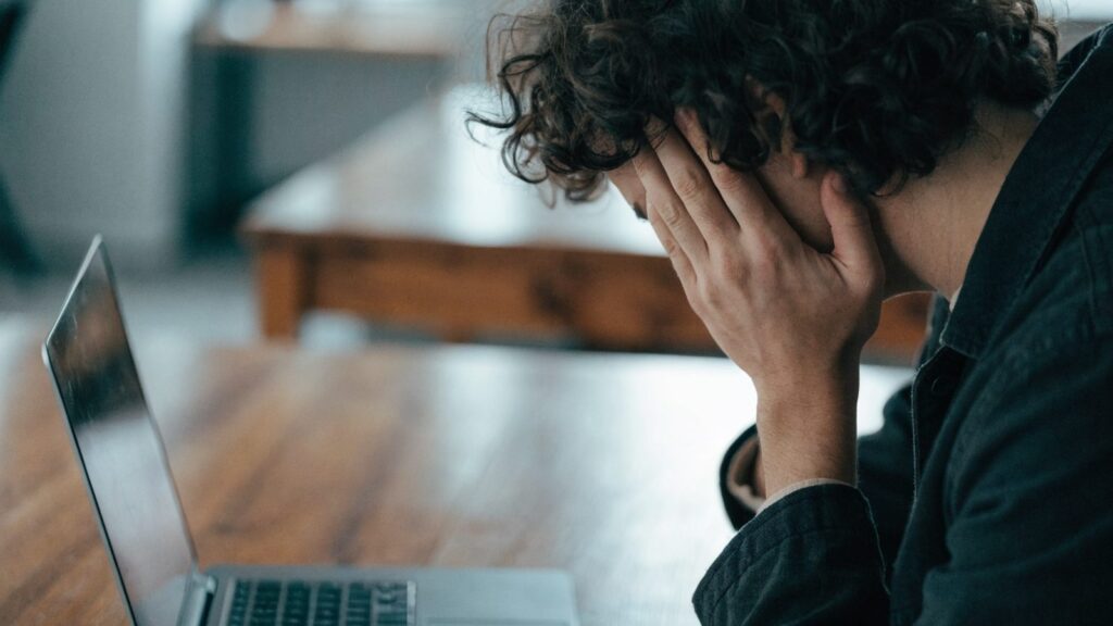 A person resting their forehead on one hand in front of a matte grey laptop on a wooden desk