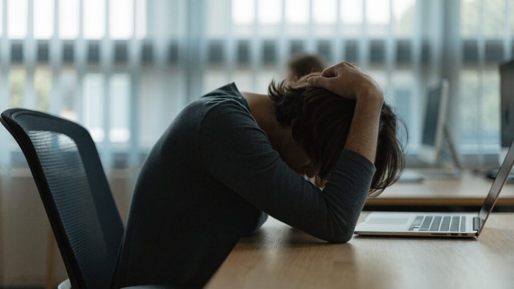 A person resting their forehead on one hand in front of a silver laptop on an office desk