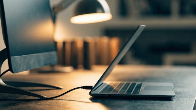 A matte grey laptop connected to a large external monitor with a blank dark screen via a cable on a workbench