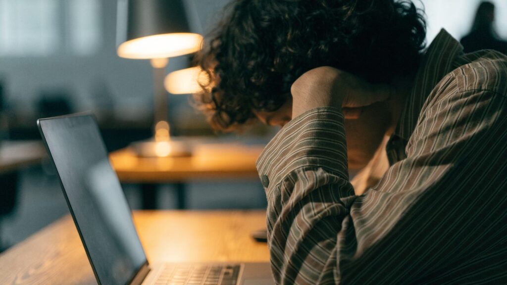 A person resting their forehead on one hand in front of a matte grey laptop on an office desk
