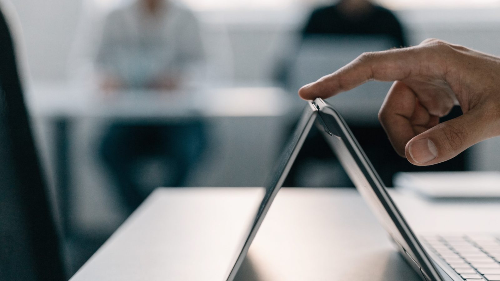 A hand touching the screen of a convertible silver laptop folded in tent mode on a white desk