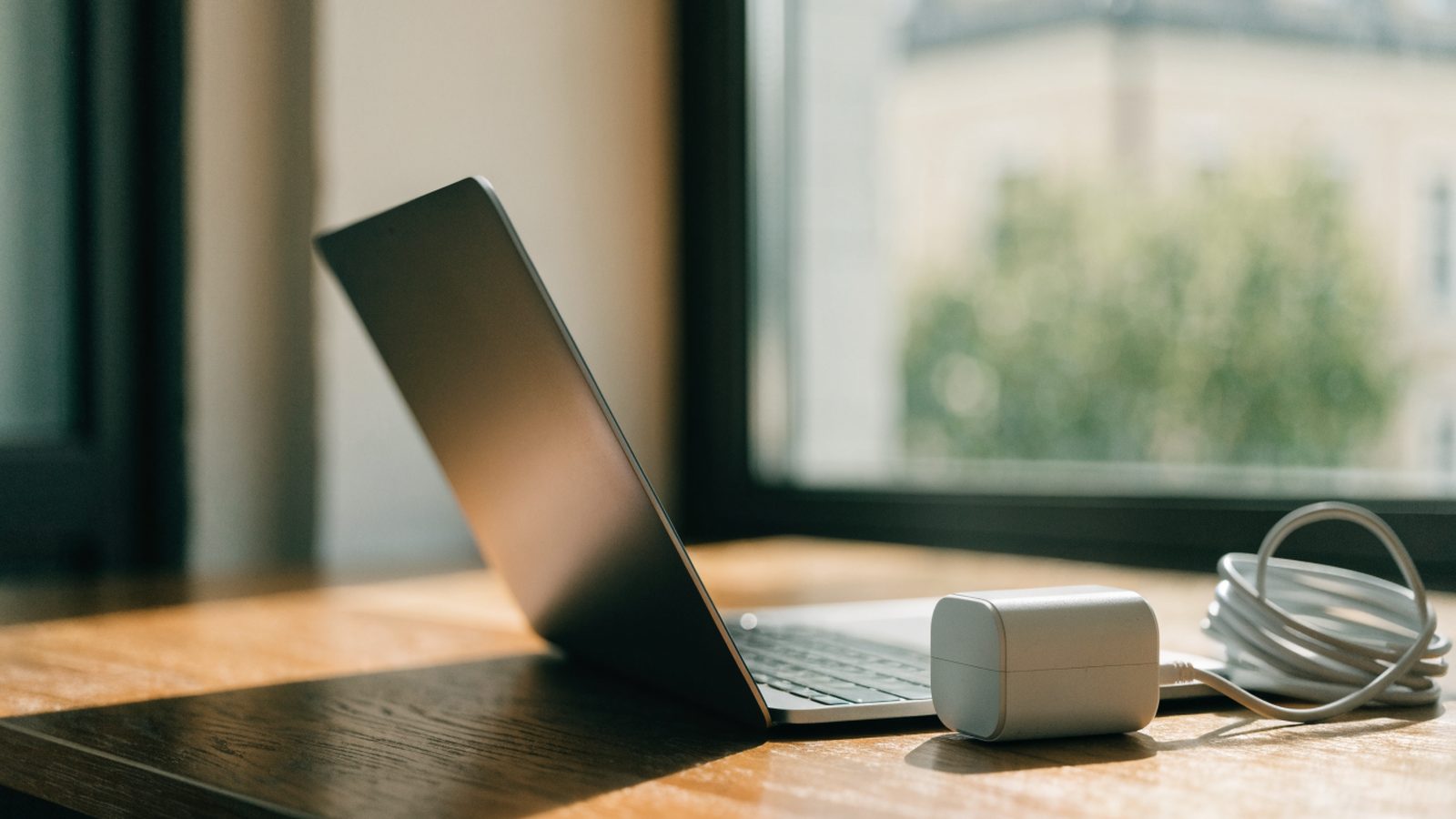 A matte grey laptop on a wooden desk next to a compact white power adapter with its cable neatly coiled