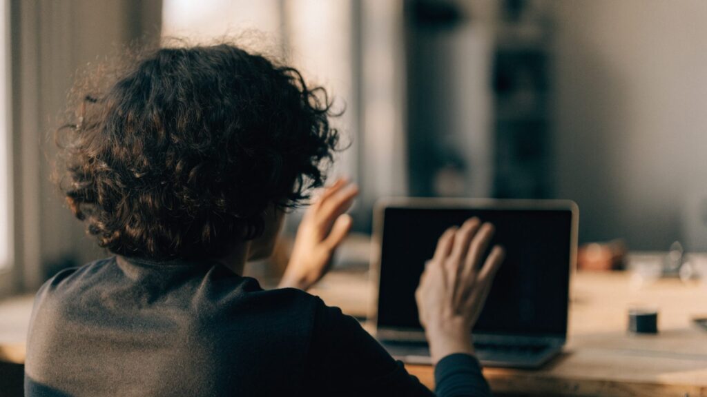 A person seen from behind with hands raised at a matte grey laptop with a dark blank screen on a workbench