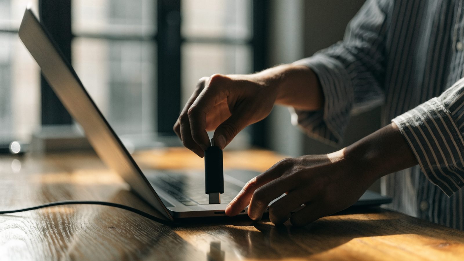 Hands unplugging a charging cable from a slim graphite black laptop on a wooden desk