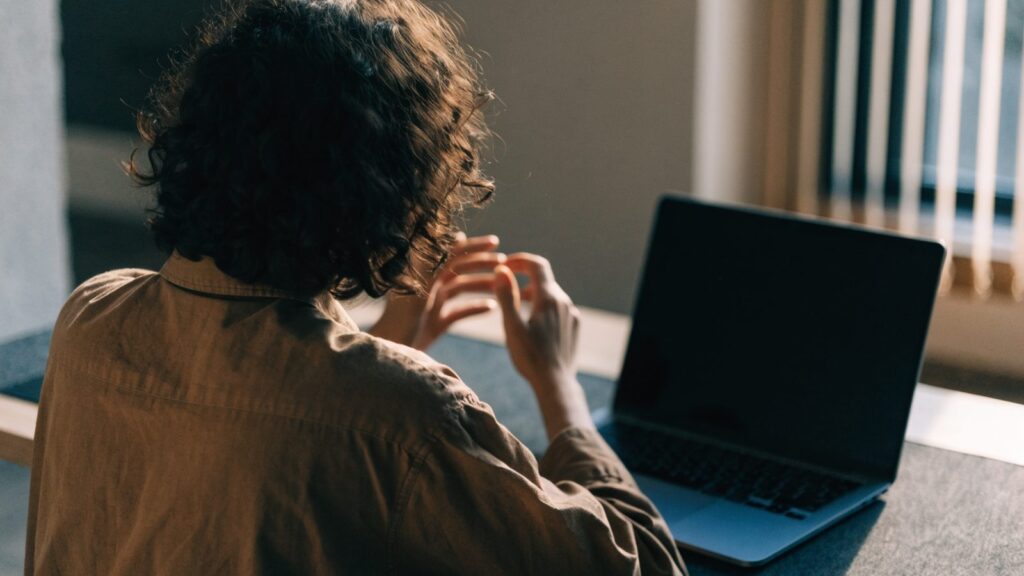 A person seen from behind with hands raised at a dark blue laptop with a dark blank screen on a dark desk mat