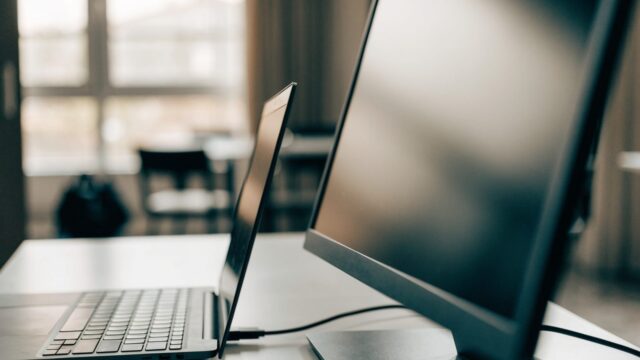 A graphite black laptop connected to a large external monitor with a blank dark screen via a cable on a white