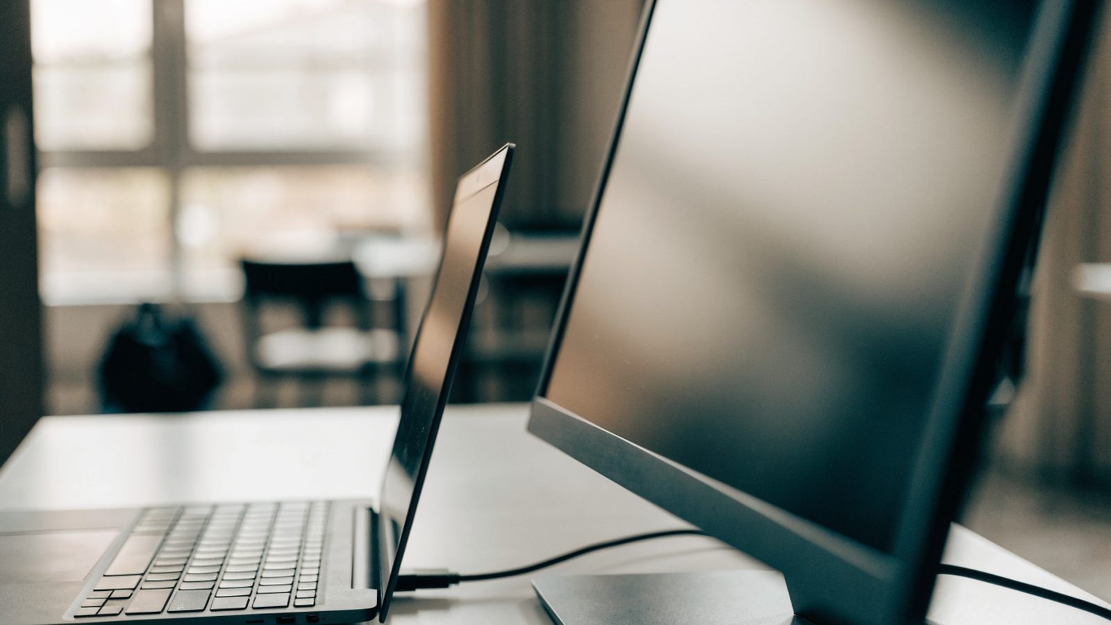 A graphite black laptop connected to a large external monitor with a blank dark screen via a cable on a white