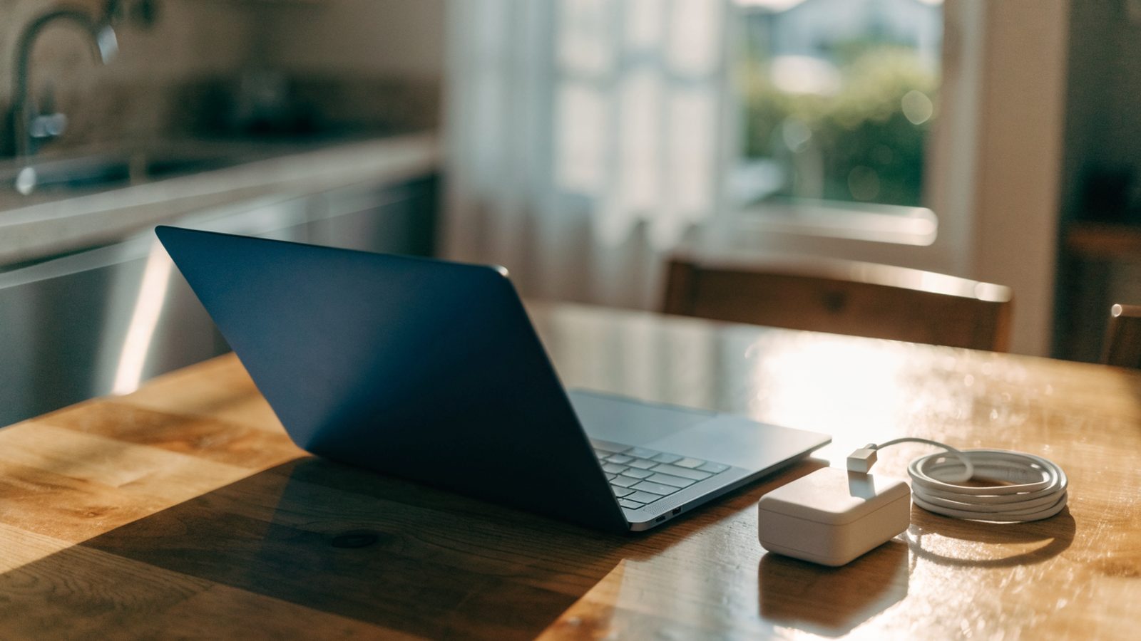 A dark blue laptop on a kitchen table next to a compact white power adapter with its cable neatly coiled