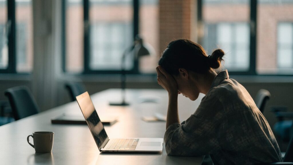 A person resting their forehead on one hand in front of a silver laptop on an office desk