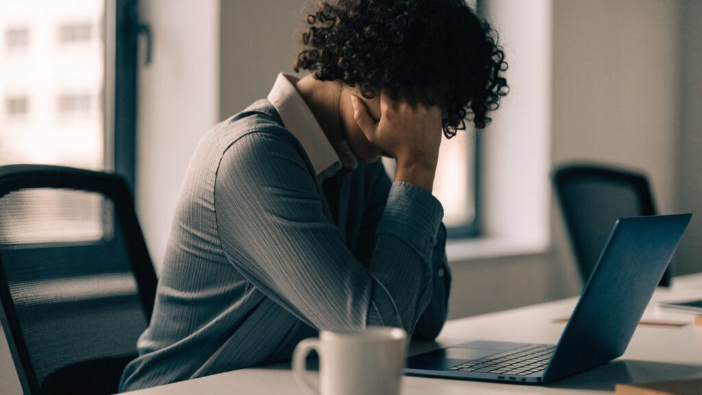 A person resting their forehead on one hand in front of a dark blue laptop on an office desk