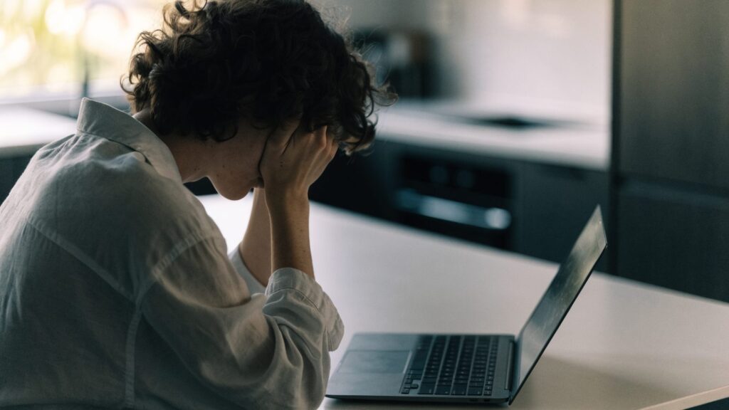 A person resting their forehead on one hand in front of a graphite black laptop on a kitchen table